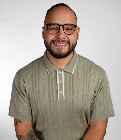 Adrian Gutierrez A smiling person with a shaved head, glasses, and a beard, wearing a ribbed green polo shirt with white trim, identified as Adrian Gutierrez, Lead Graphic Designer. Their favorite product is Beach Bum Sno.