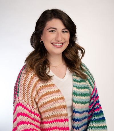 Laken Jacobs A smiling woman with long, wavy brown hair, wearing a white shirt and a colorful striped knit cardigan with shades of pink, orange, green, and blue, identified as Laken Jacobs, Digital Loyalty Specialist. Her favorite product is Pina Colada Sno with Tropic Crem