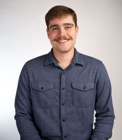 Parker Sutton A smiling man with a mustache and dark hair, wearing a dark blue long-sleeved button-up shirt, identified as Parker Sutton, Production Supervisor. His favorite product is Shark Attack Sno.
