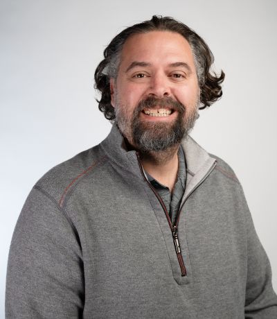 Zac Buchanan A smiling man with dark, curly hair and a beard wears a gray quarter-zip sweater with red accents, identified as Zac Buchanan, Director of Franchise Development. His favorite product is Strawberry Cheesecake Sno.