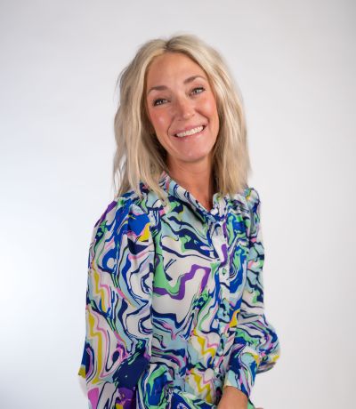 Nina Watts A smiling woman with wavy blonde hair wears a vibrant, multi-colored patterned blouse, identified as Nina Watts, Internal Operations Specialist. Her favorite product is Shark Attack Sno.