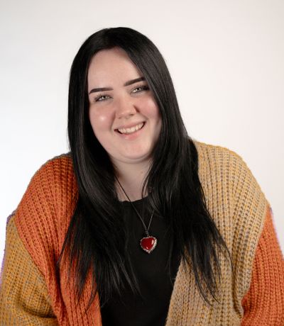 Gabby Allen A smiling person with long, dark hair wearing a multi-colored striped cardigan and a heart-shaped pendant necklace, identified as Gabby Allen, Internal Operations and Print Shop Assistant. Their favorite product is Super Sour Strawberry Sno.