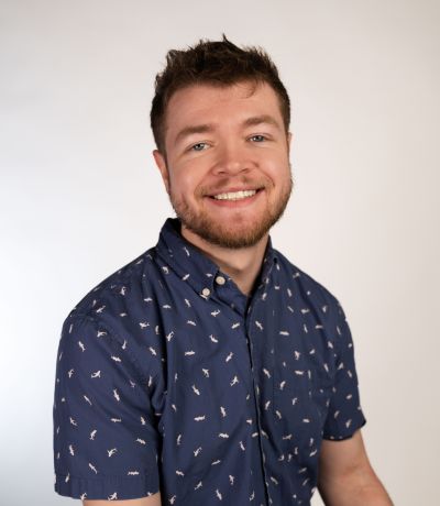 Cole Schilling A smiling man with short brown hair and a beard, wearing a dark blue short-sleeved button-up shirt with a light pattern, identified as Cole Schilling, Warehouse Specialist. His favorite product is Oasis Smoothie.